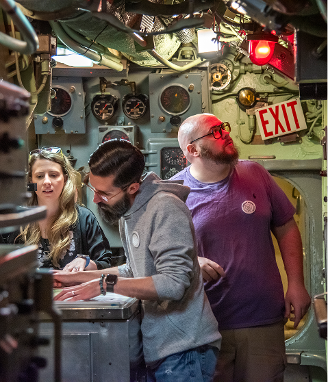 Three people inside a submarine control room with equipment and dials.