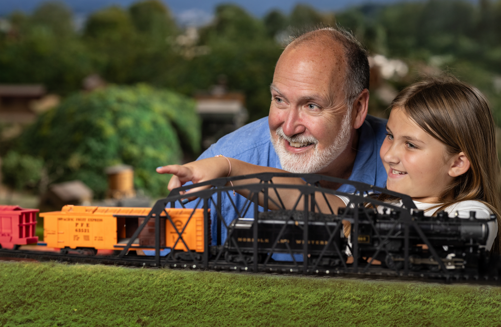 A young granddaughter looks and points out over a miniature railroad model with her grandfather.