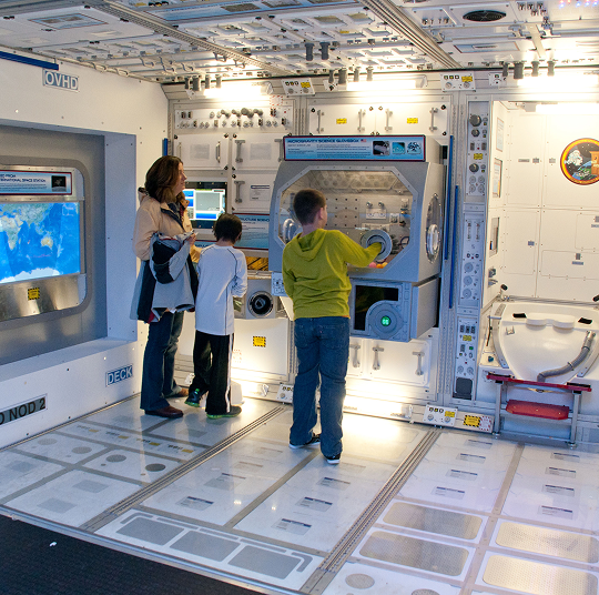 A woman and her two children interact with a space station exhibit.