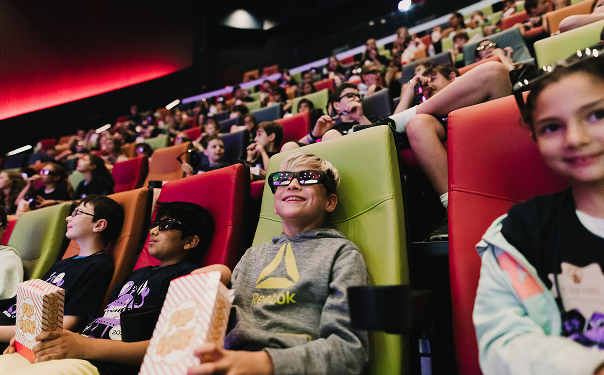 A group of children watch a movie in a theater while wearing 3D glasses and eating popcorn.