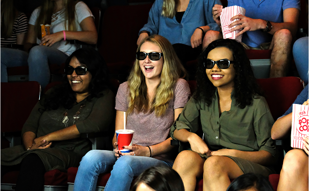 Three women wearing 3D glasses and watching a movie in a theater, with an audience in the background.