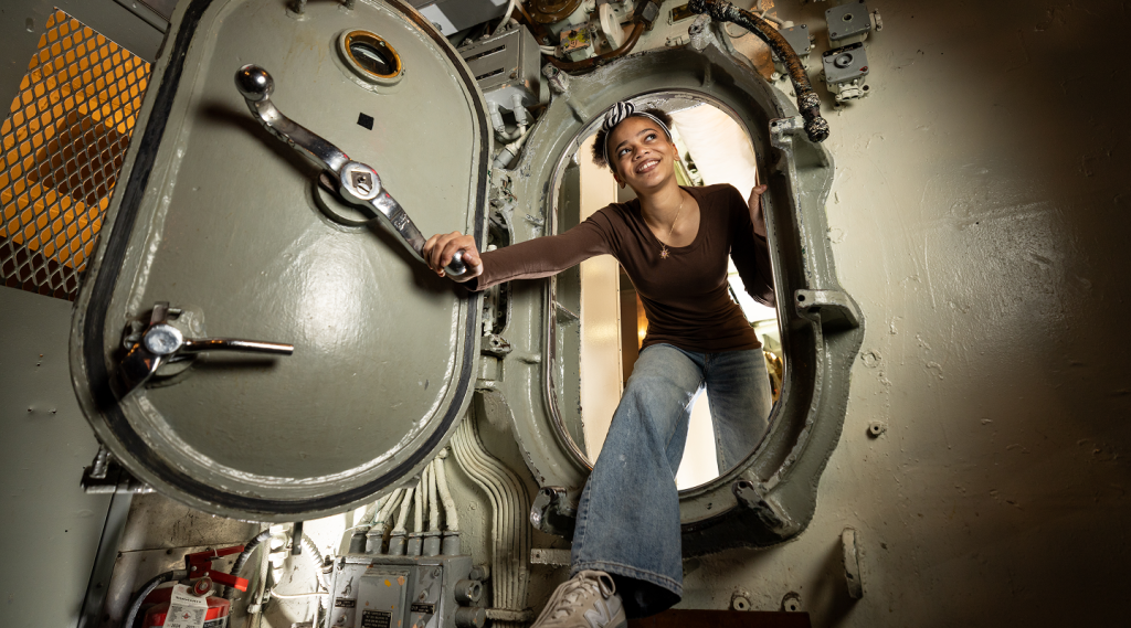 Teen girl stepping through submarine doorway.