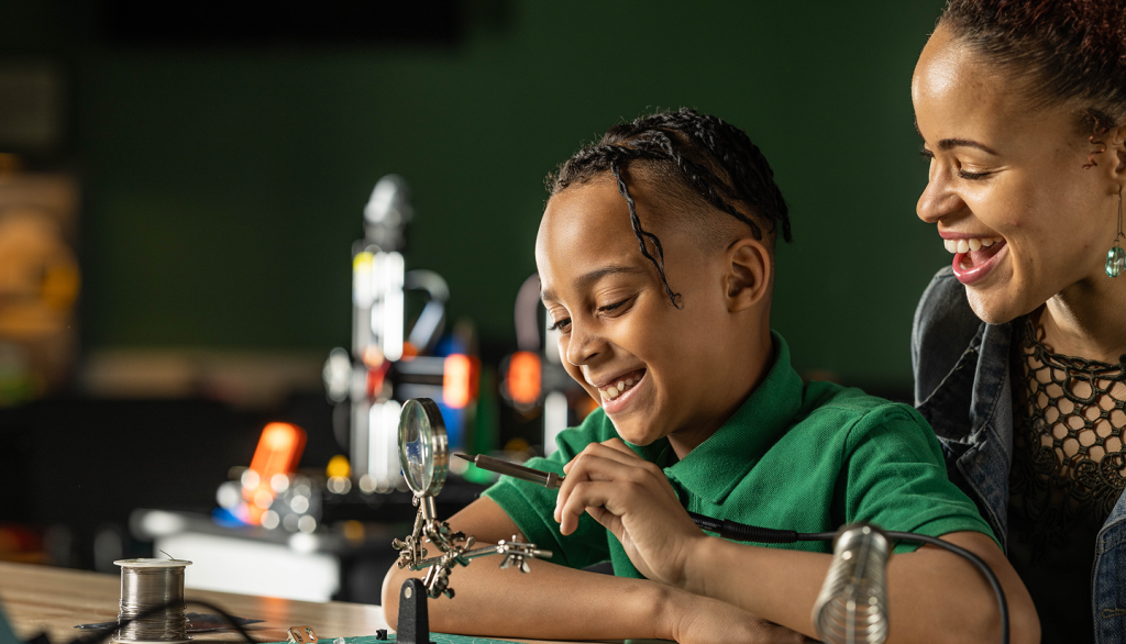 A boy and a woman smiling and working with a soldering iron and magnifying tool.