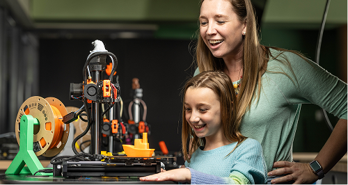Woman and girl smiling at a 3D printer with an orange boat.