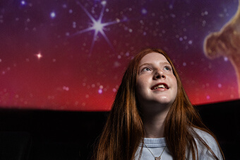A young girl looking up at a colorful, starry sky in a planetarium.