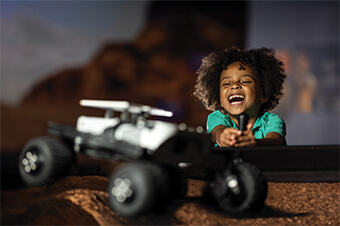 A young boy smiles while operating a remote model of a Mars Rover.
